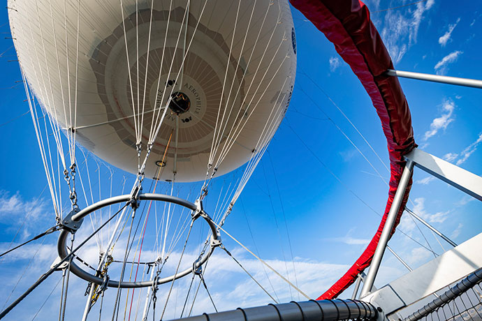 Upward view of large balloon with supporting cables and metal framework against a blue sky.