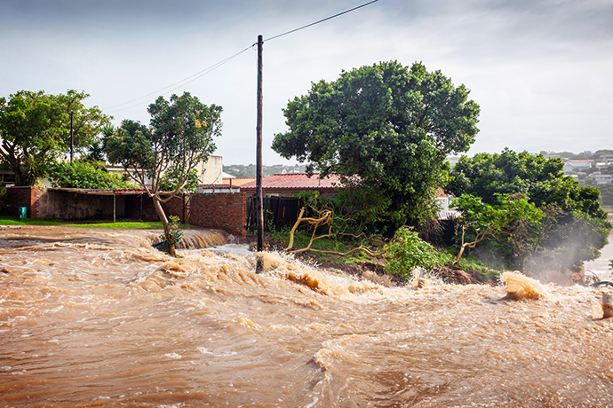 Floodwater surges through a residential area, submerging a street and rushing past houses and trees.
