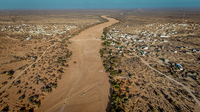 Aerial view of a dry riverbed winding through an arid landscape in East Africa, with sparse vegetation and settlements lining both sides, highlighting severe drought conditions.