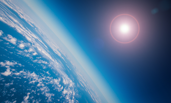 View of Earth from space, showing the planet’s curved horizon, scattered cloud cover over the ocean, and the Sun shining against a deep blue atmosphere.