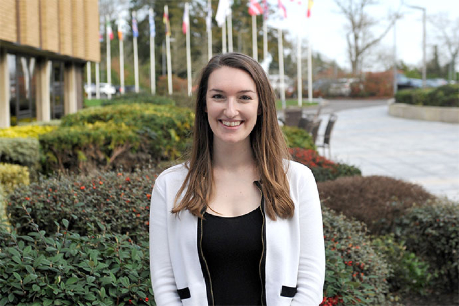 Photo of Meghan Plumridge in front of plants. In the background are flagpoles and flags.