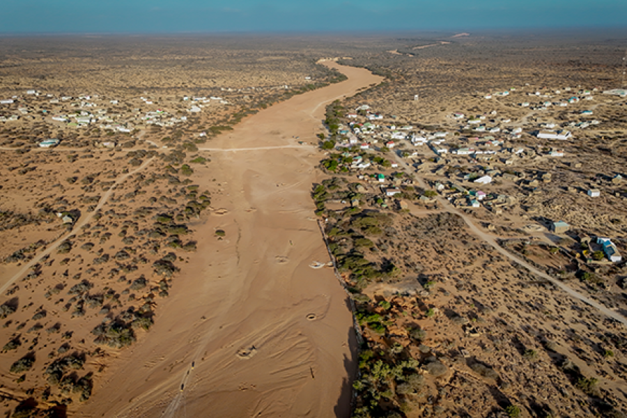 Aerial view of a dry riverbed winding through an arid landscape in East Africa, with sparse vegetation and settlements lining both sides, highlighting severe drought conditions.