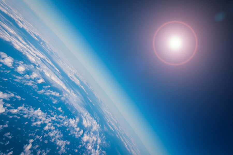 View of Earth from space, showing the planet’s curved horizon, scattered cloud cover over the ocean, and the Sun shining against a deep blue atmosphere.