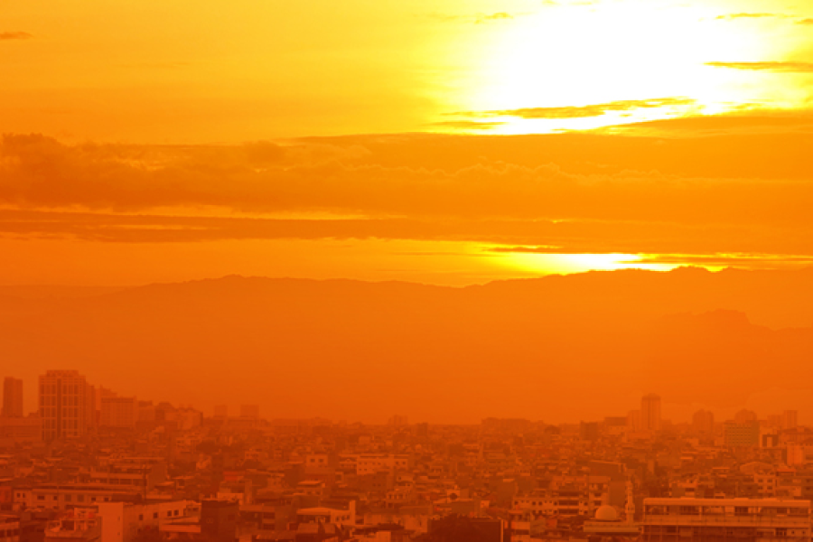 Orange-tinted city skyline at sunset during hot, hazy conditions, with dense urban buildings beneath a glowing sun and layers of atmospheric haze stretching across the horizon.