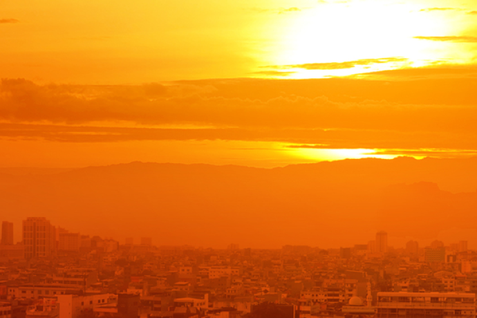 Orange-tinted city skyline at sunset during hot, hazy conditions, with dense urban buildings beneath a glowing sun and layers of atmospheric haze stretching across the horizon.