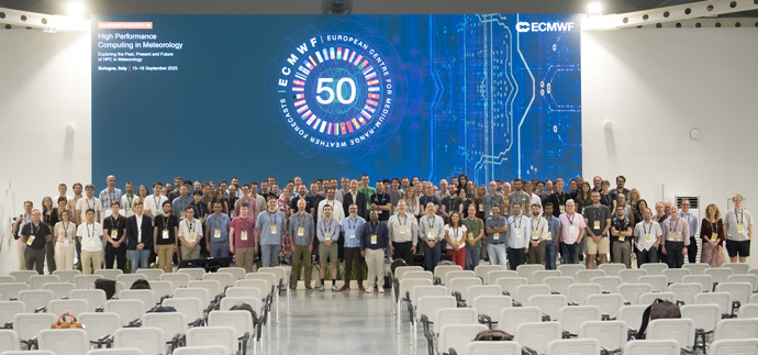 Large group of people stood behind large screen that has a blue slide with the large 50th anniversary logo on. The group are stood in front of several rows of white chairs
