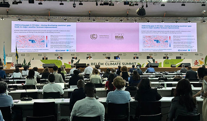 A large screen showing a presentation. Below this is a long white table with nine people sitting. An audience sits watching the presentation on several rows of white tables