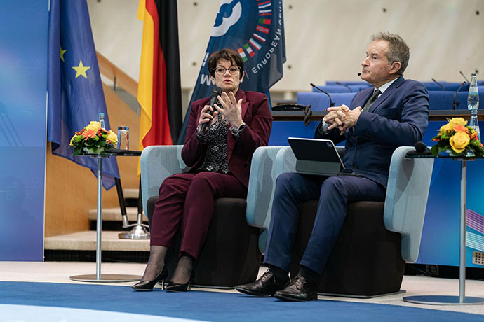 Laurence Rouil sat on a chair with a microphone speaking next to a person who has a tablet on their lap.