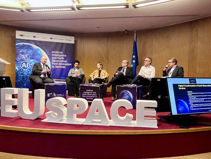 Panel of speakers seated on a stage during an EU Space event, with large “EUSPACE” letters in front, a backdrop on AI and Earth observation, and a presentation screen to the side.