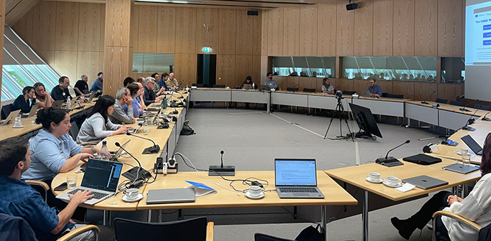 Participants seated around U-shaped conference tables during a workshop meeting, with presentations displayed on screens and laptops, microphones and cameras set up throughout the room.