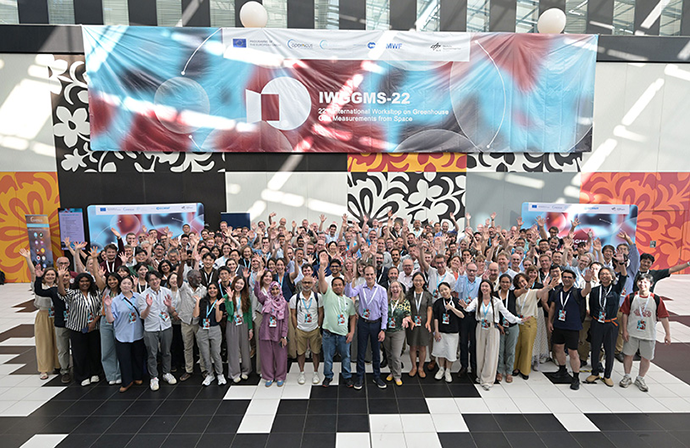 Large group photo of participants at the International Workshop on Greenhouse Gas Measurements from Space (IWSGMS-22), gathered beneath an event banner in a bright conference venue.