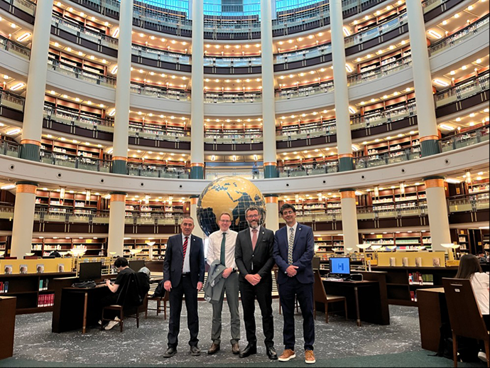 Four people in suits stood in a row in a large circular library building during a Member State visit.
