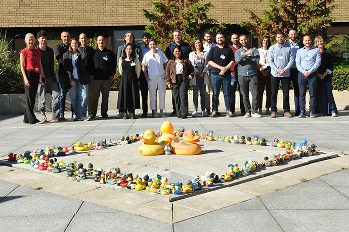 Group photo of Code for Earth participants standing outdoors behind a square arrangement of small rubber ducks on a paved courtyard, with trees and a building in the background.